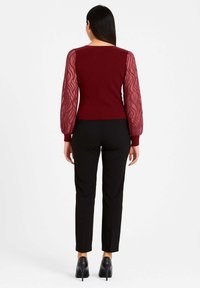 Top bordeaux in maglia con maniche a sbuffo trasparenti e testurizzate a motivo ondulato; abbinato a pantaloni neri su misura e scarpe nere con tacco alto.