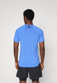 Homme de dos portant un t-shirt de sport bleu clair à manches courtes et un short noir superposé devant un fond uni.
