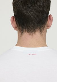 T-shirt en coton blanc avec un col rond, affichant un petit logo rouge "LEE COOPER" à l'arrière, près du col. Texture simple et lisse.