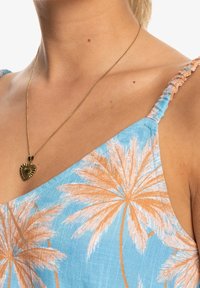 Vêtement bleu clair avec imprimé de palmiers orange. Présente des fines bretelles et une surface texturée. Porté avec un collier en pendentif en forme de cœur doré.
