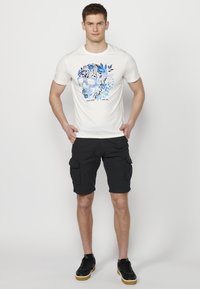 Balta trumpa rankovė t-shirt su mėlynu kaukolės grafiniu vaizdu ir gėlių motyvu. Derinama su juodais kargo šortais ir juodais sportbačiais.