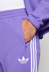 Μωβ αθλητικό μπουφάν με λευκό σχέδιο τριών ραβδώσεων, με μπροστινή τσέπη. Υφασμάτινη υφή· λευκό λογότυπο Adidas στην αριστερή πλευρά.