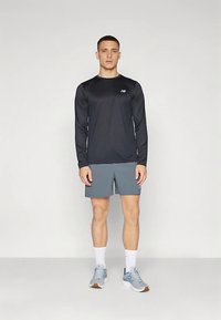 Άνδρας που στέκεται φορώντας μαύρη αθλητική μπλούζα με μακριά μανίκια, γκρίζες βερμούδες, λευκές κάλτσες και γκρίζα αθλητικά παπούτσια μπροστά από ένα απλό φόντο.