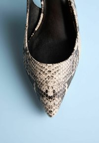 Chaussure à bout pointu en simili python blanc cassé et noir, dotée d’une doublure intérieure lisse et de côtés ajourés, posée sur un fond bleu.