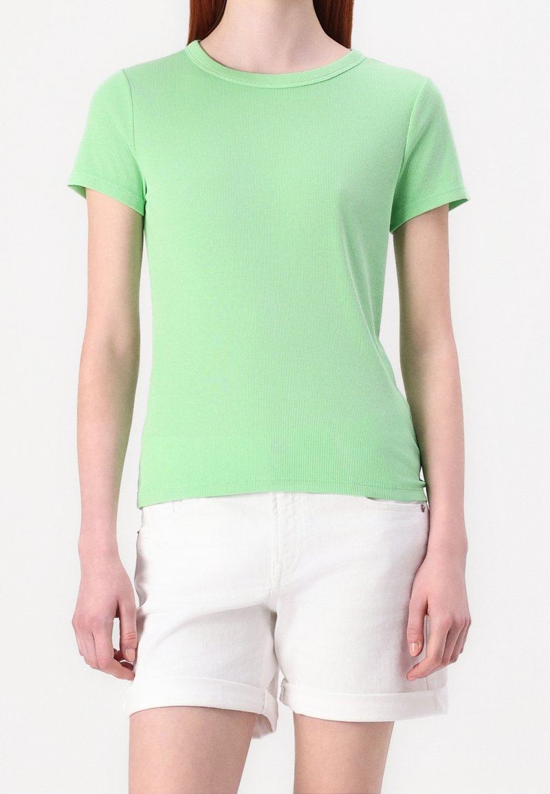 Femme portant un T-shirt côtelé vert clair à manches courtes et un short blanc avec ourlet roulé, debout devant un fond uni.