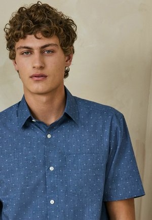 Mladý muž s kudrnatými vlasy, který má na sobě modrou košili s krátkým rukávem a jemným tečkovaným vzorem na neutrálním pozadí.