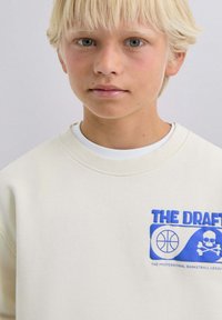 Sudadera beige clara con cuello redondo, con un gráfico azul de un logo de baloncesto y calavera, hecha de un tejido suave y texturizado.