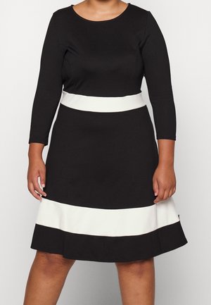 Kobieta w czarnej sukience z rękawami za łokieć i białymi poziomymi paskami na wysokości talii i na dole, stojąca na jednolitym tle.