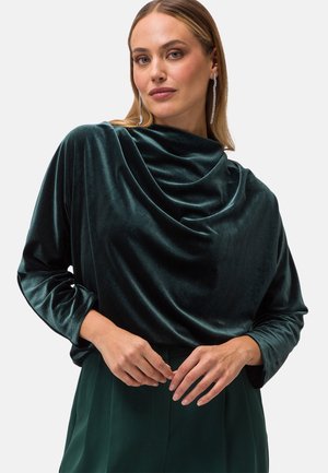Top in velluto verde con scollo drappeggiato e maniche lunghe. Il tessuto ha una consistenza liscia, riflettendo la luce in modo sottile. Design semplice, senza fantasie.