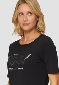 Blonde vrouw met schouderlang haar, gekleed in een zwart shirt met de tekst "Jouw potentieel kent geen grenzen," die licht naar rechts kijkt.