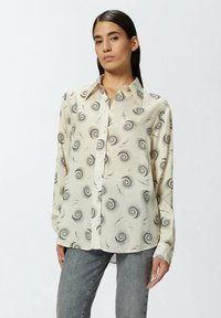 Blusa a maniche lunghe color crema con finitura lucida, caratterizzata da motivi a spirale neri e abbottonatura frontale. Tessuto morbido e leggero.