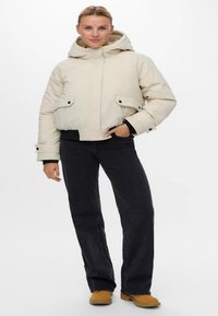 Kvinne iført en lys beige hettejakke, svarte vide jeans og brune støvler, som står med den ene hånden i lommen.