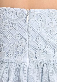 Jasnoniebieska sukienka z misternym koronkowym haftem, zamkiem błyskawicznym z tyłu oraz falbaną u dołu. Materiał wydaje się miękki i fakturowany.