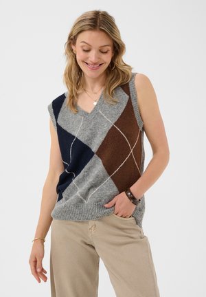 Femme souriante, portant un pull sans manches à losanges gris, marron et bleu marine avec un pantalon beige, debout devant un fond uni clair.