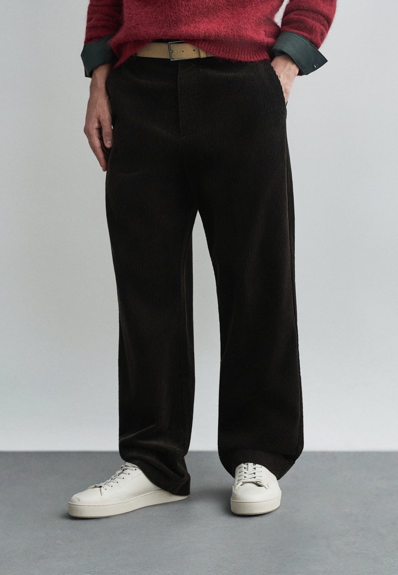 Sötétbarna kordbársony nadrág, egyenes lábszárral, bézs övvel és fehér sneakers-szel párosítva. Texturált anyag, látható bordázattal.