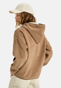 Brun fleece hoodie med en stor h�ætte, tekstureret overflade og afslappet pasform. Sammen med en lys farvet cap og beige bukser, der fremviser et afslappet look.
