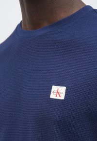 Tmavomodrý sveter s textúrou a okrúhlym výstrihom, s malou bielou štítkom s červeným logom "CK" na ľavej strane hrudníka.