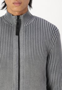 Suéter gris de crepé con cremallera y alto cuello, que presenta una cremallera negra y un tejido texturizado, mostrando rayas alternas claras y oscuras.