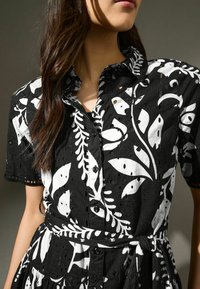 Robe chemise noir et blanc présentant un motif floral, avec des manches courtes, un devant à boutons et une ceinture en tissu. Matériau texturé avec des détails en œillets.