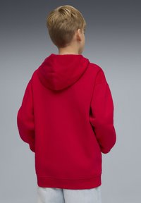 Sweat à capuche rouge à coupe ample, ourlet côtelé et grande capuche, associé à un jean de couleur claire. Le tissu semble lisse et épais.