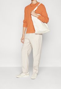 Orange tekstureret sweater, beige linned bukser og hvide sneakers med grå accenter. Bærer en hvid lærreder tote taske. Farverig perlehalskæde.