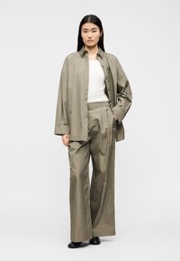 Γυναίκα που στέκεται, φοράει φαρδιά πουκάμισο σε ελιά πράσινο με κουμπιά πάνω από λευκή εσωτερική μπλούζα, ταιριαστό παντελόνι με φαρδιά πόδια και μαύρα παπούτσια, ουδέτερο φόντο.