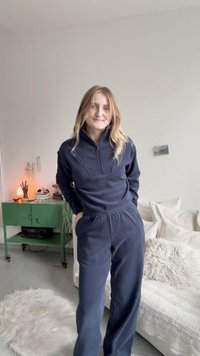 Jauna sieviete ar garām blondām matiem, tērpusies tumši zilos sporta tērpos, stāv gaišā dzīvojamā istabā ar baltu dīvānu un zaļu sānu galdu.