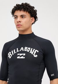 Fekete hosszú ujjú úszódressz fehér "BILLABONG" logóval, sima, rugalmas anyag, magas gallér és UV-védelem a tengerszáron.