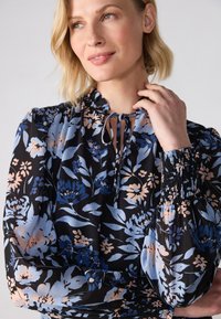 Floral blouse in black, featuring blue and peach patterns, a ruffled neckline, and smocked cuffs. Lightweight fabric with a flowing design.