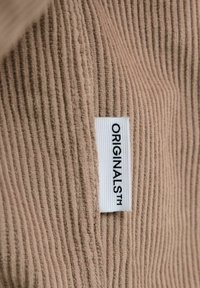 Brązowa tkanina kordurowa z pionowymi żeberkami, z białą metką z napisem "ORIGINALS™" w czarnym kolorze.