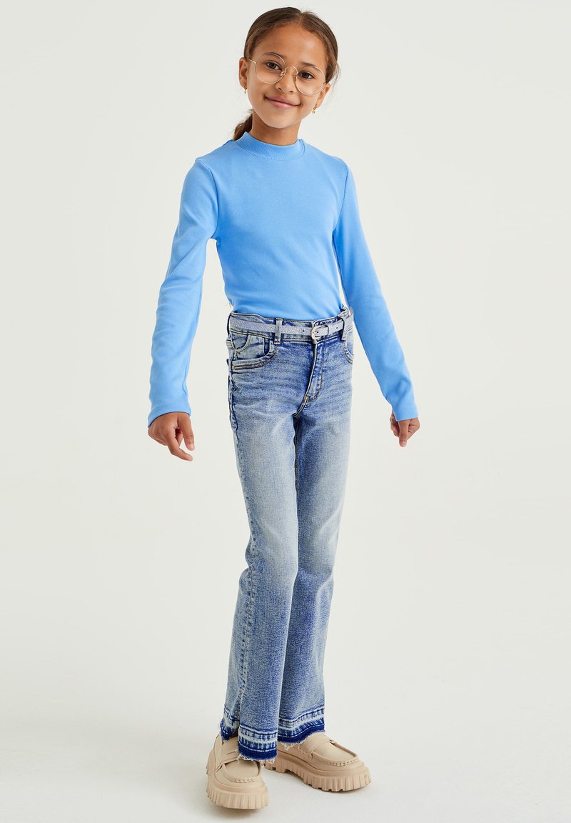 Világoskék, hosszú ujjú, testhezálló felső; világoskék, koptatott, trapéz farmernadrággal párosítva, díszített derékrésszel és mintás szegéllyel.