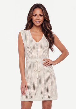 Femeie cu păr lung și ondulat, purtând o rochie bej croșetată fără mâneci, cu decolteu în V și talie cu șnur, zâmbind cu o mână pe șold.