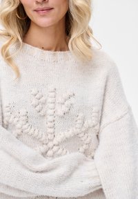 Blondynka z luźnymi lokami nosi kremowy sweter z dzianiny z wypukłym, teksturowanym wzorem kwiatowym oraz kolczyki-koła.
