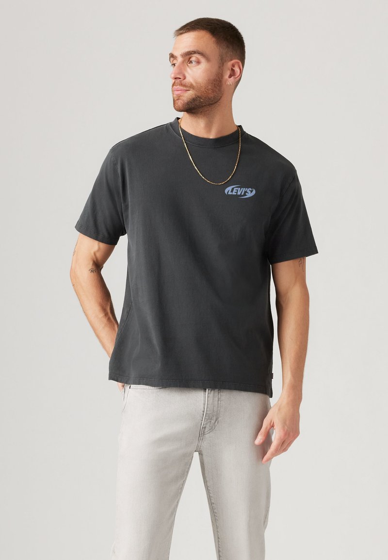 Mann mit kurzen Haaren und Bart, der ein schwarzes Levi's-T-Shirt, hellgraue Jeans und eine goldene Kette trägt, steht vor einem einfarbigen hellen Hintergrund.