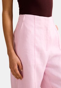 Γυναίκα που φοράει ψηλόμεσος ροζ παντελόνι από λινό με μπροστινές ραφές και πλαϊνές τσέπες, συνδυασμένο με σκούρο αμάνικο τοπ.