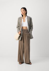 Károvaný blejzer v sivej a bielej farbe, biely crop top, hnedé nohavice s vysokým pásom a širokými nohavicami, béžová taška, otvorené čierne sandále.
