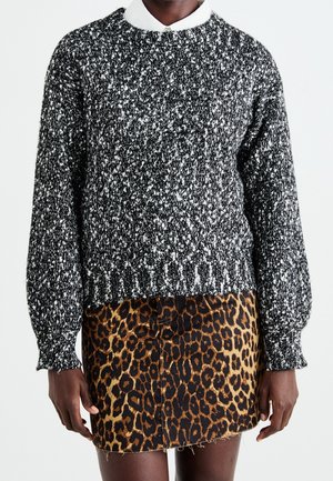 Personne portant un pull en tricot noir et blanc moucheté sur une chemise blanche à col et une mini-jupe à imprimé léopard, debout sur un fond blanc.