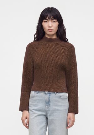Femme aux longs cheveux foncés portant un pull côtelé marron et un jean bleu clair, debout devant un fond clair uni.