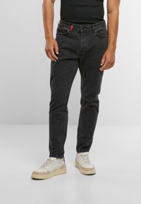 Jeans de mezclilla negra de corte Slim, con una etiqueta roja en la cintura, combinados con zapatillas blancas y beige. Textura suave, diseño clásico.