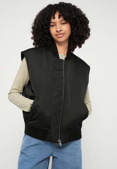 Casaco bomber preto sem mangas com fecho à frente, bainha canelada e bolsas laterais; apresenta painéis texturizados e um acabamento suave.