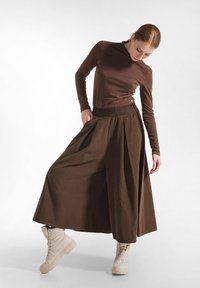 Brun langærmet bluse parret med viddebrune bukser lavet af blødt materiale, accentueret af en tætsiddende talje. Modellen har lette støvler på.