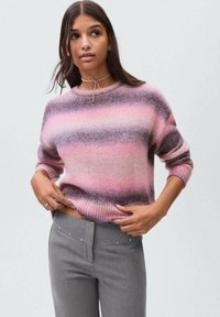 Donna che indossa un maglione a strisce rosa e viola e pantaloni grigi, mentre sistema l'orlo del suo maglione con entrambe le mani.