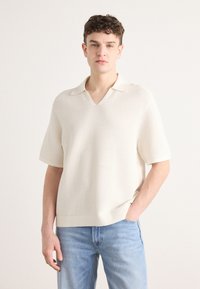 Strikket kremfarget kortermet polo med V-hals og ribbestruktur, kombinert med lyseblå denimjeans. Enkel og moderne design.