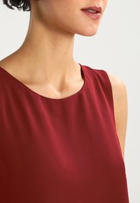 Bordeaux farvet ærmeløs bluse med en glat, satinagtig tekstur og en rund halsudskæring, der har et enkelt, elegant design.