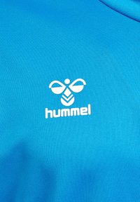 Blaues Sportshirt aus strukturierter Stoff, mit einem weißen "hummel"-Logo und einem stilisierten Emblem über dem Text.