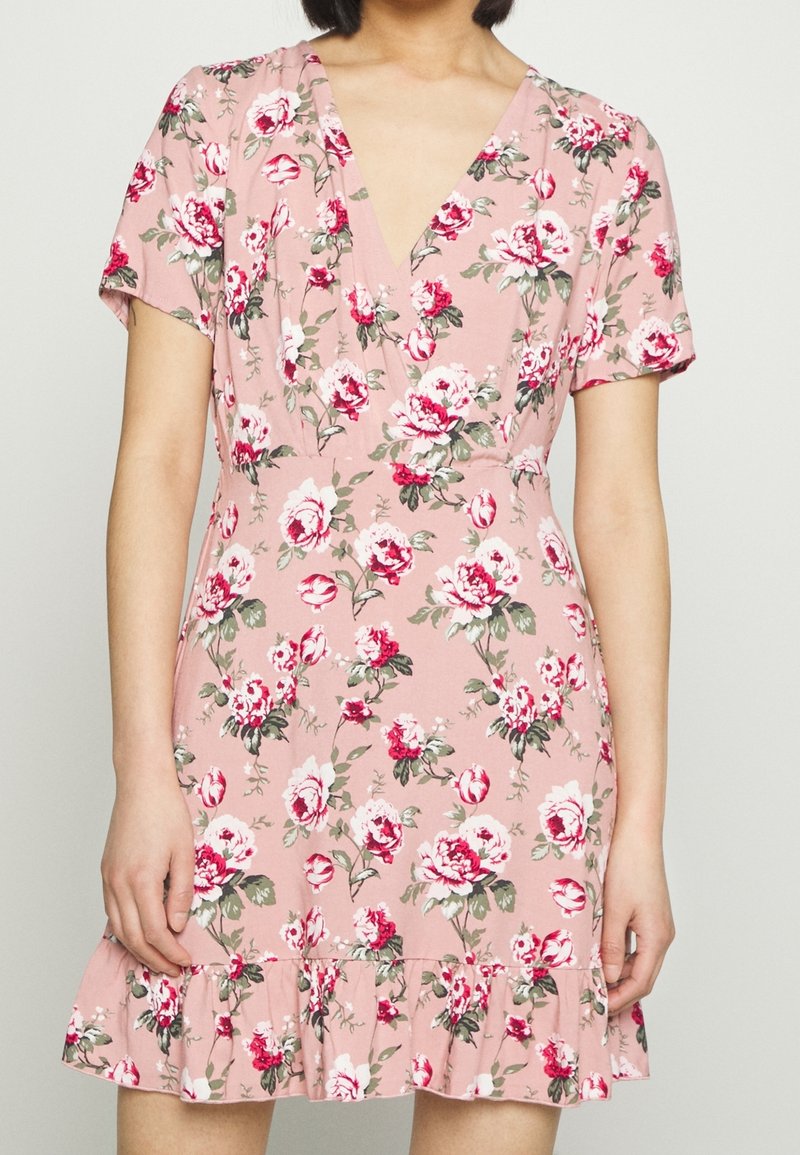 Femme portant une robe courte rose fleurie avec un col en V, des manches courtes et un ourlet à volants, debout devant un fond uni.