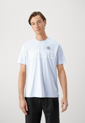 Camiseta de algodón azul claro con texto en blanco "LACOSTE" en negrita, mangas cortas, cuello redondo y un pequeño parche con el logo en el lado izquierdo del pecho.