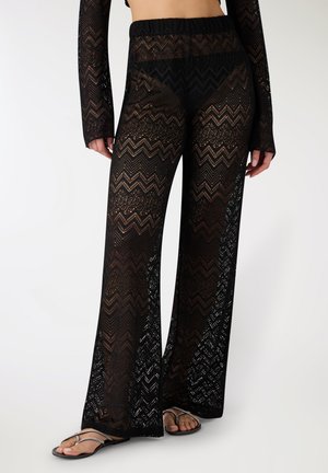 Donna che indossa pantaloni ampi neri trasparenti con motivo a zigzag e top abbinato a maniche lunghe, con sandali neri.