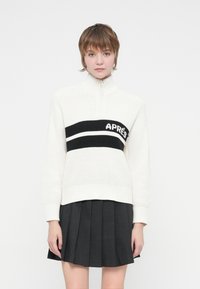 Καστόρινη ζακέτα σε off-white με ψηλό γιακά, που διαθέτει μια μαύρη ρίγα με την επιγραφή "APRÈS." Συνδυάζεται με μια μαύρη πλισέ φούστα.