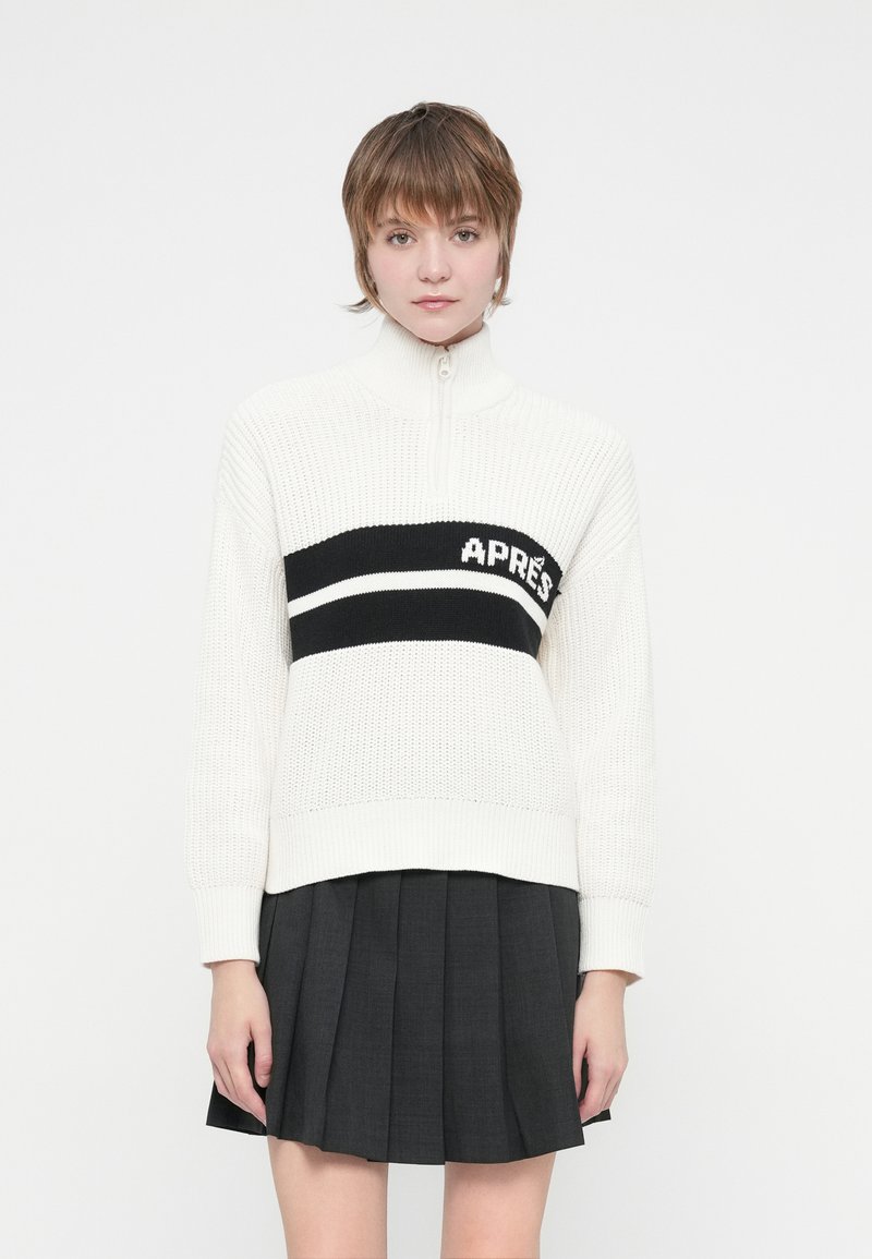 Καστόρινη ζακέτα σε off-white με ψηλό γιακά, που διαθέτει μια μαύρη ρίγα με την επιγραφή "APRÈS." Συνδυάζεται με μια μαύρη πλισέ φούστα.